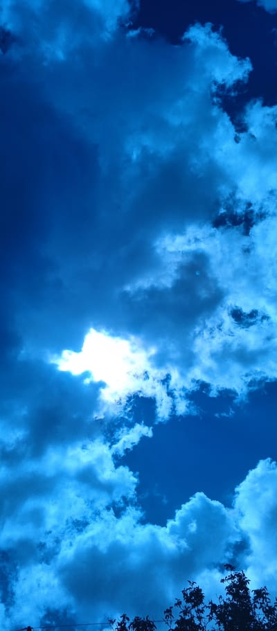 A vertical, low-angle shot captures a dramatic sky filled with billowing clouds against a deep blue backdrop. The sun is partially obscured by a large, bright white cloud formation in the upper left quadrant, casting rays that illuminate the surrounding clouds. The lower edge of the image features the dark silhouettes of tree branches and a thin wire, suggesting an outdoor scene. The overall impression is one of an impending storm or dynamic weather conditions. This scene is located in Popivka, Ukraine.