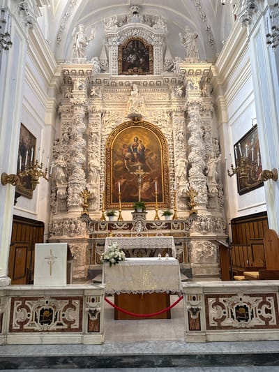 The image displays the ornate interior of a church, specifically focusing on the main altar and its surrounding apse. The style is characteristic of Baroque or Rococo architecture, featuring an abundance of intricate white stucco decorations.

At the heart of the scene is a large altarpiece painting, housed within a magnificent gilded frame. The artwork depicts a religious narrative, likely centered on the Virgin Mary and Christ, surrounded by other figures such as angels or saints, rendered with dramatic lighting and warm colors. Flanking the painting are two prominent golden candelabras, each holding multiple candles. On the altar table below, a white cloth with delicate lace trim is adorned with a vase of fresh white flowers, possibly lilies or roses, adding a touch of serene beauty.

The walls surrounding the altarpiece are a spectacle of detailed white stucco work. This includes sculpted figures of saints or angels emerging from clouds, intertwined with rich floral motifs, garlands, cherubs, and complex architectural elements like columns, pilasters, and cornices. The ceiling is vaulted and also features elaborate white ornamentation.

To the right of the altar, a simple white stand or lectern is visible, marked with the "IHS" monogram. Adjacent to this, decorative marble panels in shades of white, brown, and black display geometric and scroll patterns. One panel features a distinct central motif that appears to be a skull and crossbones, a classic memento mori symbol. A red rope barrier is also present on the right side. Overhead, chandelier-like light fixtures hang from the ceiling, and other paintings can be glimpsed on upper walls.

The bright illumination suggests daylight. No individuals are present, allowing the viewer to absorb the intricate artistry of the sacred space. This grand and historically rich setting is located in Monopoli, Italy.