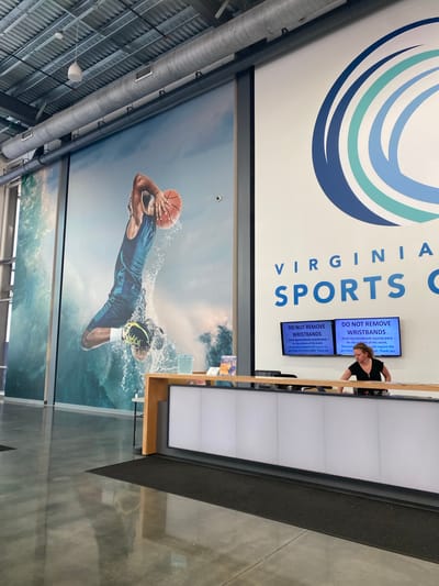 The image depicts the interior of the Virginia Sports Complex in Virginia Beach, United States of America. A large mural of a basketball player mid-dunk against a crashing wave covers the left wall. To the right, the facility's name "VIRGINIA SPORTS C" is displayed in large blue lettering, followed by a stylized blue and green logo. Below the logo, two digital screens display a "DO NOT REMOVE WRISTBANDS" message. A woman with blonde hair, wearing a black top, stands behind a reception desk, interacting with the computer system. The reception desk features a white, illuminated front panel and a light wood countertop. The floor is polished concrete, reflecting the overhead lighting and the surrounding environment. Several large industrial air ducts are visible on the ceiling.