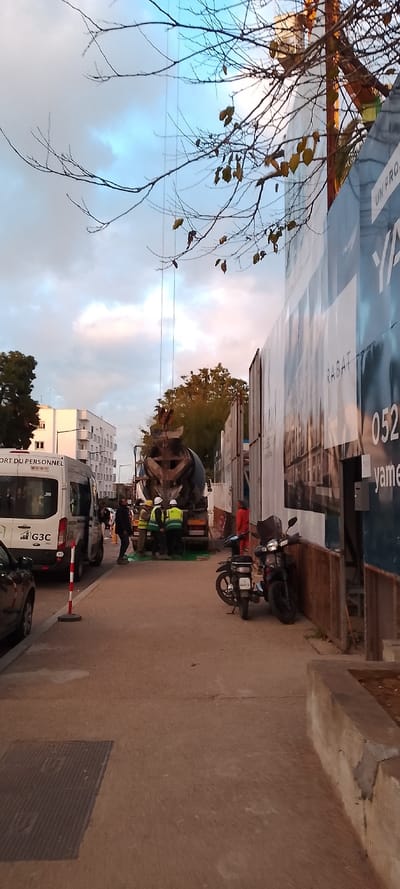 This image captures an urban construction scene during what appears to be late afternoon or early evening, marked by the warm light illuminating buildings and a partly cloudy sky with patches of blue and pink. Bare tree branches with a few lingering yellow-brown leaves frame the top of the photo.
In the center, several construction workers are engaged in an activity around a large concrete mixer truck. At least three workers are visible wearing yellow and green high-visibility vests and hard hats, with a green tarp or protective material laid on the ground beneath the mixer. To their left, a white commercial van, labeled "TRANSPORT DU PERSONNEL" (personnel transport) and bearing a "G3C" logo, is parked, alongside a partially visible dark car and a red and white traffic cone.
On the right, a tall construction hoarding or fence lines the street, adorned with large advertisements. Visible text on these ads includes "RABAT", a partial phone number "052", and fragments like "UN PRO..." and "YAMEN". Two motorcycles are parked upright against this hoarding. In the background on the left, a white, multi-story apartment building stands under the soft light. The foreground features a concrete sidewalk with a metal manhole cover on the left and a low concrete wall or planter on the right. The scene suggests ongoing construction work in a built-up area.