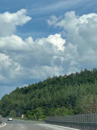 The image depicts a highway scene with a dense forest covering a hillside on the right side of the road. The sky above is partly cloudy with dramatic formations of white and gray clouds against a blue sky. A silver car is driving on the highway, heading away from the viewer. The highway has multiple lanes and is bordered by a sound barrier on the right. A circular traffic sign with a red border and a horizontal bar is visible near the edge of the road, indicating a prohibition. The landscape appears to be rural or semi-rural, with no visible buildings or other urban structures. The scene is set during daylight hours.