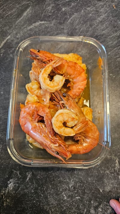 An overhead shot captures a clear, rectangular food container filled with cooked shrimp and a side dish, set against a dark, mottled background. The container, likely made of glass or plastic, holds numerous shrimp in various states of preparation. Several large, bright orange-red shrimp with their heads and shells intact are prominently displayed, characterized by their curled bodies and distinct antennae. Interspersed among these are smaller, shelled shrimp, revealing their lighter, opaque pink flesh.
A golden-brown sauce or liquid is visible at the bottom of the container, enveloping a yellowish-orange food item that forms the base of the meal. This base could be mashed potatoes, polenta, or a similar soft accompaniment. The container rests on a dark, textured surface, possibly a granite or marble countertop, which exhibits subtle grey and white striations. In the upper right corner, a small portion of a human hand, including fingertips and a thumb, is visible, suggesting interaction with the container, perhaps holding or placing it. The image depicts a prepared meal, ready for consumption or storage, with focus on the vibrant colors and fresh appearance of the seafood. No text is visible in the image. The setting appears to be an indoor environment, such as a kitchen or dining area.