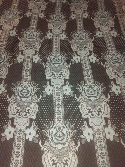 The image displays a close-up, overhead view of a patterned fabric, likely a textile or blanket, with a floral and ornate design. The dominant colors are brown and a light cream or beige. The pattern features repeating vertical bands with floral motifs at the top and bottom, interspersed with smaller floral elements and a dotted background. There are no people or discernible environmental elements that would place this image in a specific city or location like Haveli Jhangranwali, Pakistan. The photograph focuses solely on the texture and design of the fabric.