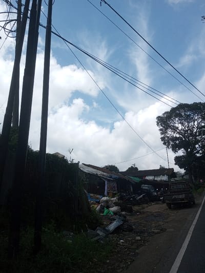 The image depicts a roadside scene in Ura-Dozhi, Bhutan, under a blue sky with scattered white clouds. On the left, three dark, vertical utility poles rise from the foreground, supporting multiple black electrical wires that extend diagonally across the upper frame. Below the poles, a dense mass of dark green foliage covers an unseen structure or embankment. To the right of the foliage, a collection of informal structures with corrugated metal and tarp roofs lines the roadside. In front of these structures, piles of white and green bags, along with discarded items including what appears to be a broken television screen, are visible on the uneven ground. A dark pickup truck and several motorcycles are parked off the paved road, which features a white edge line and runs diagonally from the bottom right. In the background, traditional white-walled buildings with dark roofs are partially obscured by a large, leafy tree on the right side of the frame. No individuals are visible in the scene.