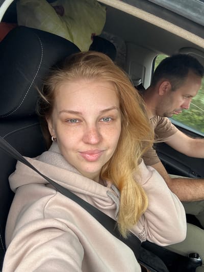 The image shows two people in a car. A woman in the foreground, wearing a light pink hooded sweatshirt and a seatbelt, is looking at the camera. She has blonde hair with hints of red, light skin with freckles, and green eyes. She is wearing hoop earrings. Behind her and to the right, a man with short brown hair and a brown t-shirt is driving. He is wearing a seatbelt. The interior of the car appears to be black with white stitching on the seats. Outside the car, greenery is visible, suggesting a rural or suburban setting. The image was taken in Anteplevo (Okhotinskoe S/P), Russia.