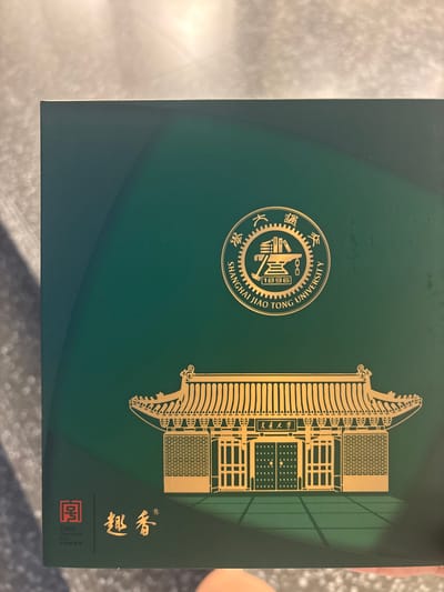The image displays the cover of a dark green box with gold embellishments. At the top center is a circular emblem that reads "SHANGHAI JIAO TONG UNIVERSITY 1896" in English, surrounded by Chinese characters. Below the emblem is a depiction of a traditional Chinese building with a tiered roof, intricate doorways, and a brick facade, all rendered in gold. In the bottom left corner, there is a red square symbol with Chinese characters, and next to it, more gold Chinese characters and a registered trademark symbol. The background of the box features subtle, wavy lines in a slightly lighter shade of green. The image is taken from a slight angle, with a portion of a person's hand visible at the bottom edge. The scene appears to be a close-up product shot, likely taken indoors. The location is Minhang District, Shanghai, China.
