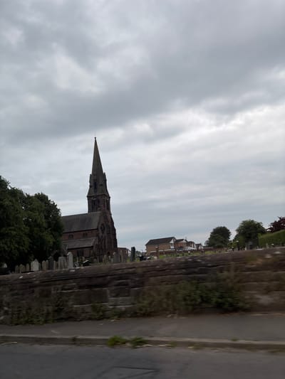 The image shows a view of St. John's Church in Winsford, United Kingdom, on a cloudy day. The church, made of red brick, has a tall, pointed spire. In front of the church, a cemetery is visible with numerous gravestones and a low stone wall. Behind the church, several houses can be seen. The foreground of the image is a blurry view of a road and pavement.
