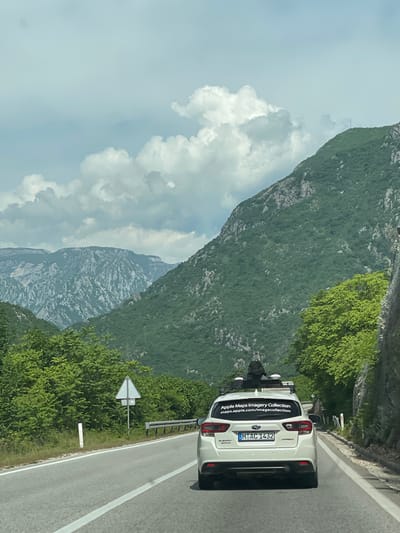 A white Subaru Impreza with the license plate "MO AC 1432" is parked on the right side of a paved road. The car features an "Apple Maps Imagery Collection" sticker on the back. The road runs alongside a lush, green mountain. The sky is visible with scattered clouds. The location is in the vicinity of Donji Jasenjani, Bosnia and Herzegovina, based on the probable location of Apple Maps imagery collection. The view is seen from another vehicle, indicating the photo was taken by someone traveling on the road.