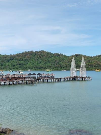 The image depicts a waterfront scene in Layeun, Indonesia, featuring a wooden pier extending into the ocean. The pier has small, thatched-roof pavilions along one section and culminates in a structure resembling twin towers, constructed from white, open-framework material. In the background, a forested hillside provides a backdrop to the calm, light-blue water. Scattered boats are visible on the water's surface. The sky is partly cloudy. In the lower foreground, rocks sit at the edge of the shoreline.