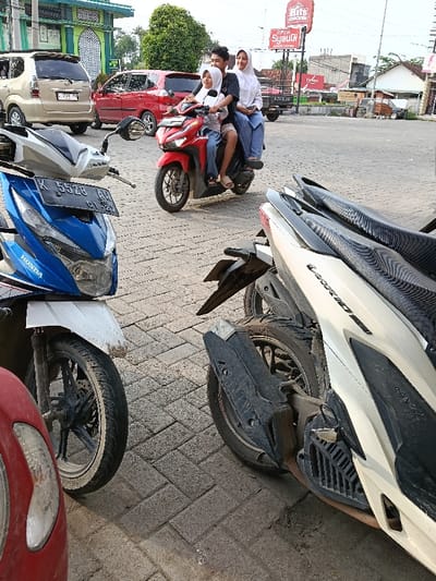 The image depicts a street scene in Sumberejo, Indonesia, under daylight conditions. The foreground shows grey interlocking brick pavement and three motorcycles. On the far left, a portion of a red motorcycle is visible. Centrally, a blue and white Honda scooter (license plate K 5528 AV 01-27) is parked. To its right, the rear section of a white/cream Honda Vario 150 scooter is visible, featuring a textured black seat cover and a black exhaust guard.

In the midground, a red Honda scooter is in motion, traveling from left to right across the frame. It is occupied by three individuals. The driver is a male with short dark hair, wearing a black t-shirt and beige shorts. In front of the driver, a female child is seated, wearing a white long-sleeved shirt, blue pants, and a white head covering. Behind the driver, a female passenger is seated, also wearing a white long-sleeved shirt, blue pants, and a white head covering.

Behind the moving scooter, a gold/beige minivan (license plate 7541 AV) and a red car are visible. Further back, a black flatbed truck is partially seen. The background on the left features a green building with arched window designs. On the right, commercial structures are visible, along with a prominent red sign displaying "Bits Chicken" and "Optik Syukur."