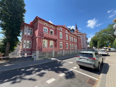 A light-green Toyota RAV4 hybrid is parked on a street in Międzyzdroje, Poland, on a sunny day. The car is facing away from the camera, with its rear end visible. The license plate reads "PZ 25810". In the background, a large, ornate red building with white trim and balconies stands beside a tall tree. The building has a steep roof and multiple windows. The street is paved and has white lane markings. A white car is parked further down the street, and pedestrians are visible on the sidewalk. The sky is blue with a few scattered clouds. The scene appears to be in a town or resort area, likely during daytime given the bright sunlight and clear sky.