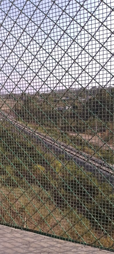 The image captures a view through a green chain-link fence, which occupies the majority of the frame and is the most prominent feature. The fence has a diamond-shaped mesh pattern, and its individual wires appear somewhat thick and coated in a dull green color, with some lighter green reflections or accents.

Through the fence, a landscape unfolds, indicating the photograph was taken from an elevated position, possibly a bridge or an overpass. At the top of the image, above the landscape, a clear, light blue sky is visible.

The middle and background reveal a verdant, hilly, and somewhat rural or undeveloped area. Lush green foliage and trees cover the slopes. Scattered within this greenery, particularly towards the center and upper right, are a few distant buildings or structures, suggesting a small settlement or houses nestled among the hills.

A railway track is a significant feature in the midground and lower-midground, curving through the landscape from the left side of the frame towards the lower right. The track is laid on a bed of gravel (ballast), and the wooden or concrete sleepers are visible beneath the steel rails. Further to the far left, a structure resembling a railway bridge or overpass can be partially seen.

At the very bottom of the image, a concrete or paved surface is visible, likely the platform or structure from which the fence rises. Shadows of the fence's mesh pattern are distinctly cast onto this surface, indicating bright sunlight and suggesting it is daytime. The overall impression is one of looking down upon a scenic, semi-rural environment on a clear, sunny day. No text is visible in the image.