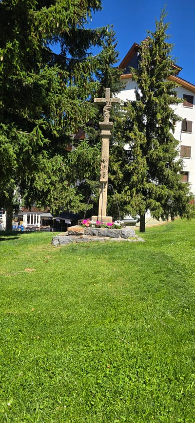 This media file depicts a stone wayside cross situated in a grassy area adjacent to a building, with numerous evergreen trees framing the scene. The cross itself is ornate, featuring carvings on its upright and horizontal beams, and appears weathered. It is positioned on a stone base adorned with a small arrangement of colorful flowers, predominantly pink and white.

In the background, a white building with brown shutters and a dark-tiled roof is visible, suggesting a residential or institutional structure. The surrounding trees are dense and green, creating a natural, somewhat enclosed atmosphere around the cross. Beyond the trees, hints of other buildings and infrastructure, including tables and chairs, suggest the presence of a village or town.

The activity or event taking place is not explicitly shown, but the setting implies a peaceful outdoor environment, possibly a memorial site or a landmark within a community. The bright sunlight and clear blue sky indicate a pleasant, sunny day. The overall mood is serene and perhaps a little solemn due to the presence of the cross. There is no visible text in the image. The location context suggests Sallent de Gállego, Spain.