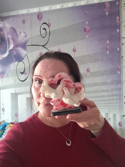 A middle-aged woman with brown hair is captured in a close-up, likely a selfie, shot indoors in Сусады-Эбалак, Russia. She wears a plain red long-sleeved top and a silver necklace with a circular pendant. Her right hand, raised to her face, holds a decorative figurine. This figurine features two white doves nestled within a translucent pink heart-shaped structure adorned with smaller red heart details, all resting on a black base. The figurine partially obscures the woman's nose and mouth, but her left eye is visible, looking directly at the camera with a neutral to slightly engaged expression.
The background is dominated by a patterned wall covering, possibly wallpaper or a tapestry. The design includes a large, soft purple rose or flower on the left side. Abstract elements such as dark, swirling lines and horizontal greyish-white stripes also feature. Numerous spherical purple and pink elements, resembling hanging beads or bubbles, are scattered across the background, contributing to a decorative aesthetic. In the bottom left corner, a small portion of a brightly colored, possibly knitted or woven item, such as a blanket or throw, is visible. The lighting is bright and even, consistent with an indoor daytime setting. There is no visible text.