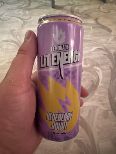 A hand holds a can of "LIT ENERGY" beverage. The can is purple and yellow, with the words "LEMONADE," "LIT ENERGY," and "BLUEBERRY DONUT" printed on it. The background appears to be a textured surface, possibly a tablecloth. There is no indication of the city of Abay, Kazakhstan in the image.