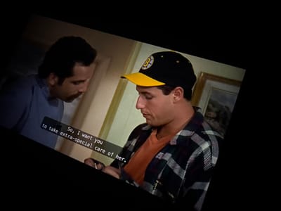 This image depicts an indoor scene in Hallandale Beach, United States, featuring two adult males. The man on the left, with dark hair and a mustache, wears a light blue collared shirt and is observed looking downwards towards the man on the right. The man on the right wears a black baseball cap with a yellow brim and a yellow Boston Bruins 'B' logo, an orange t-shirt, and a dark plaid flannel shirt. He is looking downwards at an object held in his hands, which are partially visible at the bottom center. Closed captions are overlaid on the lower left of the image, displaying the text: "So, I want you to take extra-special care of her." The background consists of a plain, light-colored wall with a framed picture, possibly a landscape, hanging on the right side. The scene is illuminated by an unseen light source, suggesting an interior setting. The interaction between the two individuals appears to be a focused exchange, possibly involving instruction or a transfer of an item.