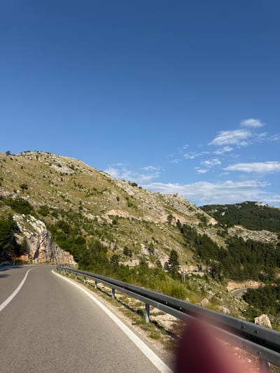 An asphalt road with white lane markings curves through a mountainous landscape under a clear blue sky with scattered white clouds. A metal guardrail lines the right side of the road. The mountainside to the right is covered in sparse green vegetation, rocks, and trees, with a winding road visible further up the slope. The mountainside to the left features rocky cliffs and more vegetation. The scene appears to be in a rural, mountainous area. The image was taken in Bukovica, Montenegro.