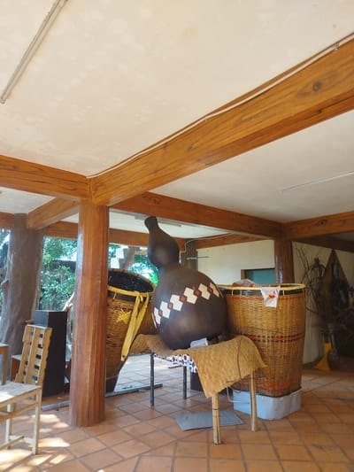 The image depicts an interior space in An Phu Ward, Vietnam. The room is characterized by a terracotta tiled floor and a ceiling with visible wooden beams. Several large woven baskets are present, one of which contains a large, dark-colored gourd-shaped object adorned with geometric white patterns. The gourd rests on a small table covered with a patterned cloth. Additional wooden chairs and objects are visible in the background, and the open-air setting allows for a view of greenery outside. The scene appears to showcase local craft or cultural objects within a covered but open-air structure.