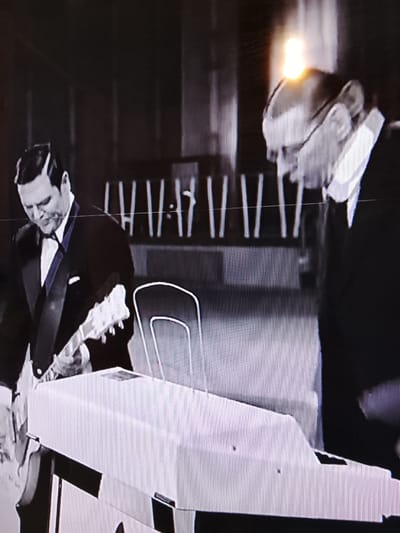 A black and white image features two men in suits performing music. The man on the left holds a guitar and looks down. The man on the right wears glasses. In front of them is a keyboard. Behind them is a row of vertical objects. The location, Высокиничи, Russia, is not apparent in the image.
