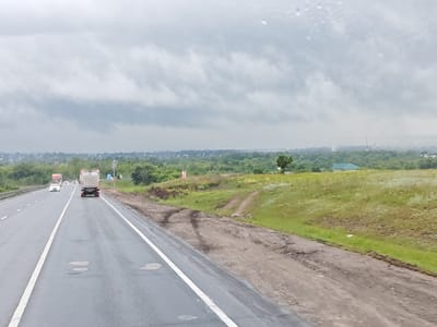 The image captures a highway scene on an overcast day, with a heavy gray sky dominating the upper portion of the frame. The road, which appears wet, is a multi-lane asphalt surface marked with white lines. In the foreground, a large gray truck with its brake lights illuminated is traveling in the right lane. Further ahead in the left lane, a white passenger car is visible. To the left of the highway, a red truck is also in motion.
The roadside environment features a gently sloping grassy embankment on the right, with patches of disturbed soil and some sparse vegetation. A single tree stands on the embankment in the middle distance. Beyond the embankment, the landscape opens up to reveal a sprawling green expanse with scattered trees and in the far distance, a settlement with buildings, including a house with a green roof and what appears to be an industrial facility with tall chimneys. The overall impression is a rural or semi-rural setting on the outskirts of a town, possibly Chubovka, Russia, given the context. The weather suggests a possibility of rain or recent precipitation.