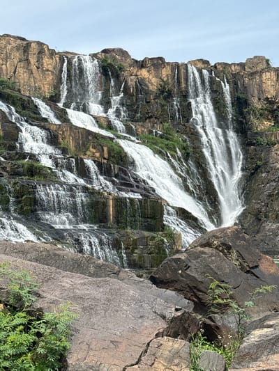 The image displays the Pongour Waterfall in Tân Phú, Vietnam. The waterfall cascades down a series of rocky tiers, creating a powerful and dynamic spectacle. The rocks are rugged and varied in color, with patches of green vegetation clinging to their surfaces. The water is a bright white, contrasting sharply with the dark rock. The sky above is a pale blue, with some wispy clouds. In the foreground, large boulders are visible, covered in moss and small plants. The overall impression is one of natural beauty and raw power.