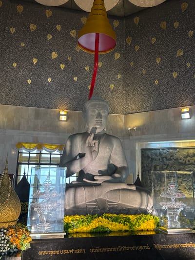 The image depicts the interior of a shrine with a large stone statue of Buddha at its center. The Buddha statue is seated in a meditative posture with its hands raised in a mudra. The shrine's ceiling is adorned with a mosaic of small dark tiles and gold leaf patterns resembling leaves. A large yellow bell-shaped object hangs from the ceiling above the Buddha, with a red pole extending downwards. The walls behind the Buddha are covered in light-colored marble. In front of the Buddha, there are elaborate floral arrangements of yellow and green flowers, along with a clear glass case containing a crystalline artifact. To the left and right of the Buddha, there are ornate golden spire-like objects. A carved relief depicting figures is visible on the wall to the right of the Buddha. Below the Buddha statue, an inscription in Thai script is present on a dark surface. The scene appears to be within a temple or sacred site, likely in Chang Khoeng, Thailand, given the context provided.