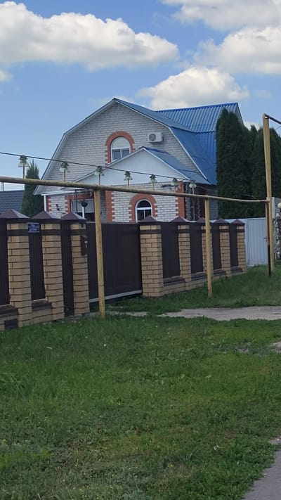 In the city of Aidarovo, Russia, a brick house with a blue metal roof stands behind a decorative brick fence with dark brown metal inserts. The house features white brickwork with orange brick accents around two arched windows, one of which is visible on the upper floor. An air conditioning unit is mounted on the exterior wall. The fence is topped with pointed caps. A metal gate is visible within the fence. A yellow metal pole, part of an electrical line support, is positioned in front of the fence. The foreground consists of a grassy area with some weeds, and a paved path is visible at the bottom right. The sky above is blue with scattered white clouds.
