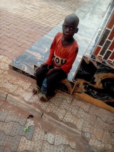 A young child is seated outdoors, looking directly at the viewer with a serious expression. The child has dark skin, closely cropped or shaved dark hair, and dark eyes. They are wearing an orange long-sleeved shirt that appears somewhat worn, with a white and black graphic on the front that reads "YOU." The child also wears dark pants and light green, open-toed sandals. A clear plastic water bottle with a blue cap is held between their legs.
The child is sitting on a low, tiled ledge or step. The tiles on this ledge and an adjacent structure are dark, featuring a swirling pattern of black, gray, and possibly white or beige. Behind the child and to their right, another part of this structure rises, topped with reddish-brown, brick-like textured tiles.
The ground in front of the child and extending into the foreground is paved with interlocking concrete pavers. These pavers are light brown or beige and feature a repetitive star-shaped pattern. The ground appears somewhat dusty or covered with light dirt in areas. There is a small piece of green debris and a thin white stick or straw visible on the paving stones in the bottom left corner of the image. The overall lighting suggests it is daytime.