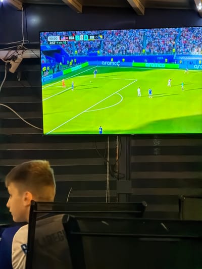 In Nević Polje, Bosnia and Herzegovina, a young boy with light brown hair is seated, facing to the left of the frame. He is wearing a white t-shirt with blue accents. In the background, a large television screen displays a soccer match. The scoreboard shows "90:00" and the score as "USA 2 - 0 BIH". The field is green, with players in blue and white uniforms visible. The stadium seating is packed with spectators in various colors. The overall environment appears to be an indoor space with dark, striped blinds covering the walls. Electrical cords are visible hanging from the ceiling on the left side of the image.