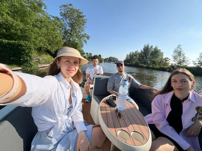 This is a horizontal, eye-level, outdoor, medium-shot photograph taken from the perspective of someone in a boat on a canal, looking towards the front and right. The scene is set in Rhenoy, Netherlands, on a sunny day with a clear blue sky.

In the foreground, on the left, a woman wearing a straw hat with a pink ribbon, a white open-button shirt tied at the waist, and a blue and white striped dress is holding up a phone to take a selfie. She is smiling and looking at the camera. She wears silver hoop earrings and a silver ring on her left hand.

Behind her, two men are seated. The man closest to the camera is wearing a grey t-shirt, a baseball cap, and shorts. He is looking ahead. Beside him, slightly further back, another man in a white t-shirt and shorts is also looking forward, holding a steering wheel.

To the right of the frame, a woman with long dark hair, wearing a pink striped open-button shirt over a black top, is seated and looking at the camera with a neutral expression. She is holding a small teddy bear charm on her right arm and wears silver hoop earrings and a silver ring on her right hand.

A wooden table with two cup holders is situated between the two seated women. On the table are a bottle of water, a smartphone, and a pair of sunglasses.

The boat is on a calm canal, with green trees and vegetation lining the banks. In the distance, more trees and some buildings are visible under the clear blue sky. The overall atmosphere suggests a leisurely boat trip on a warm day.