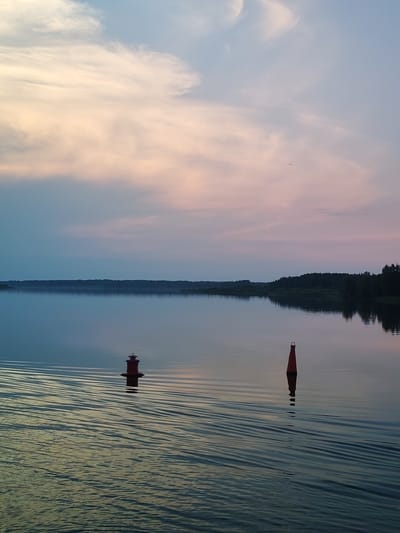 The image depicts a tranquil natural scene at dusk, likely on a body of water near Ul'yanovo, Russia. The sky is a gradient of soft blues and pinks, with wispy clouds and a faint crescent moon visible. The water is calm, reflecting the colors of the sky and creating gentle ripples. Two buoys are present: a red conical buoy with a handle on top, and a red cylindrical buoy with a light fixture. Both are floating on the water. In the distance, a dark, tree-lined shoreline is visible, with the trees appearing as a silhouette against the fading light. The overall atmosphere is serene and peaceful.