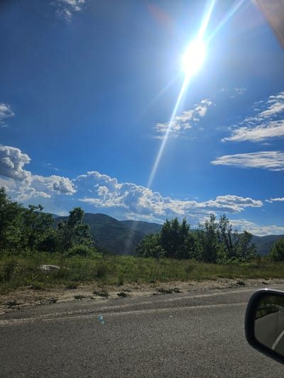 The image depicts a view from a vehicle moving along a paved road, likely in a rural area near Liaskovo, Bulgaria. The sky is blue with scattered cumulus and cirrus clouds. The sun is visible, producing a bright glare. To the right of the road, there is a verge of grass and trees leading to a hillside or mountain range in the distance. The side mirror of the vehicle is visible in the top right corner. There are no people visible. The main subjects are the sky, clouds, landscape, and road. The interaction is the movement of the vehicle through the landscape. The environment suggests a sunny day in a mountainous or hilly region.