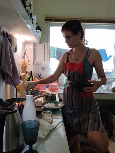 A woman is standing in a kitchen, possibly in Ban Patan, Thailand, and appears to be preparing food. She is wearing a multicolored striped apron over a dark tank top and has short, dark hair. In her right hand, she holds a dark plate. Her left hand is reaching towards a cooking pan on the stove. On the counter, there are various kitchen items including a white bottle, a glass, a kettle, and containers of food. The kitchen appears to be small and functional, with shelves above the counter holding more supplies. A window is visible in the background, showing what looks like laundry hanging outside. The lighting in the scene is bright, casting some shadows.