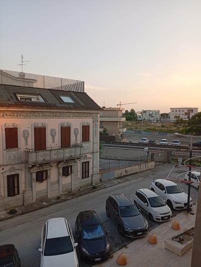 The image shows a street scene at dusk in Civitanova Marche, Italy. The foreground features several parked cars, including a white sedan, a dark blue hatchback, a gray SUV, and two white SUVs further down the street. A balcony with ornate detailing extends from the facade of a pale pink, multi-story building on the left side of the frame. The building has brown shuttered windows and architectural embellishments.
Behind the building, a railway line runs horizontally across the middle ground, with a concrete barrier alongside it. Beyond the railway, there are more buildings, including modern apartment blocks and a construction crane visible in the distance. The sky is a gradient of pale blue and soft orange, indicating the setting sun. The environment appears to be a mixed-use urban area with residential buildings, infrastructure, and open spaces. There are no people visible in the image.