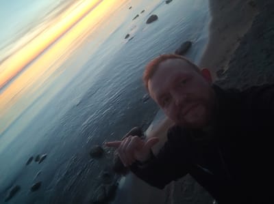 The image shows a selfie taken outdoors during what appears to be sunrise or sunset, as indicated by the bright orange and yellow hues in the sky reflecting on the water. The scene is dominated by a body of water, likely a lake or sea, with several rocks visible both in the water and on the shoreline. A man with red hair and a beard is in the foreground, holding his right hand up, pointing towards the water with his index finger. He is wearing a dark shirt. The image has a diagonal orientation, with the sky and water filling most of the frame. The location is identified as Motornoe, Russia.