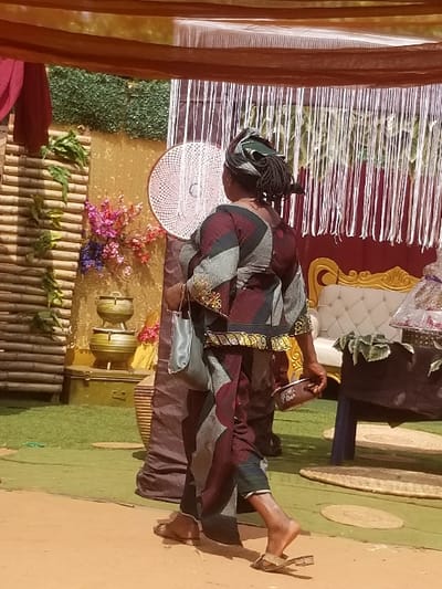 In Ochadam, Nigeria, an adult individual, viewed from the back, walks from left to right across a decorated outdoor event setting. The person, with dark skin and dark dreadlocks partially covered by a patterned green and dark headwrap, wears a traditional two-piece garment in maroon, grey, and black geometric patterns, featuring a golden patterned waistband. A light grey clutch bag hangs from their left arm, and a small, rectangular, brown container is held in their right hand. Light-colored open-toed slippers are worn on their feet, with the right slipper appearing partially dislodged.
The immediate foreground is a light brown, possibly earthen surface, transitioning to a green carpet or artificial turf further back. Overhead, brown and orange fabric drapes form a canopy. The decorated backdrop features, from left to right: a maroon fabric panel; a vertical bamboo slat structure with green foliage; a golden-toned wall adorned with clusters of pink, purple, and yellow artificial flowers. Below the flowers, two stacked golden, lidded vessels rest on a dark base, with a dark green trunk-like object visible beneath. A large white, circular, woven decorative element is centrally affixed to the golden wall. To the right, a vertical screen of thin white strings hangs in front of a dark red fabric. Further right, a section of a cream-colored tufted sofa with golden ornate trim is visible, alongside a dark blue/black table draped with green leaves, and a circular woven mat on the green ground.