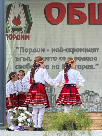 A group of four young girls in traditional Bulgarian folk costumes are standing on a stage in front of a large banner. The banner features a logo with a flame symbol and text in Cyrillic, including "ОБЩИНА ПОРАИМ" (Municipality of Pordim). The girls are wearing white blouses with embroidered details, red pleated skirts with horizontal striped patterns, and headbands adorned with colorful pom-poms and ribbons. The two girls in the foreground are dancing, with their hands on their hips, while the two girls behind them appear to be observing. A bouquet of white flowers is placed on the edge of the stage in front of the girls. The background banner also depicts an architectural illustration of a building, possibly in Pordim. The setting appears to be an outdoor event.