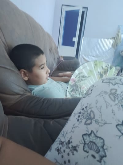 In Tor-Tor mahalla, Uzbekistan, a young boy with dark hair is reclining on a plush, brown armchair. He is wearing a light blue t-shirt and is looking to his right, with his head propped on his arm. A decorative fan, adorned with an intricate floral and bird design, is positioned behind him and to his right. In the foreground, a person is partially visible, wearing patterned pajama pants with a white base and a blue floral design. The background shows a partially open door with a blue pane, and corrugated metal sheeting. The lighting suggests an indoor setting, possibly during the day.