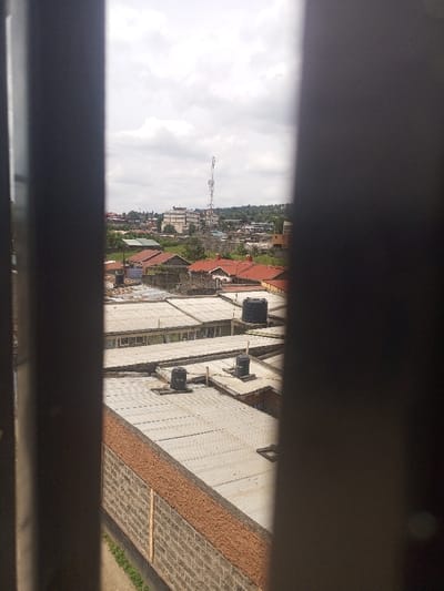 The image depicts an elevated urban landscape in Kiamaina ward, Kenya, viewed through two dark, vertical foreground bars. A grey concrete block wall, topped with a reddish-brown textured aggregate material, forms the immediate midground, with a narrow strip of grass and dirt path at its base. Behind this wall, a series of low-rise buildings with corrugated metal roofs are visible, several featuring black plastic water tanks. In the background, additional residential structures with both red-tiled and corrugated roofs are interspersed with green foliage. A prominent, tall lattice telecommunications tower rises centrally above the distant buildings. Several multi-story buildings, including a lighter-colored commercial-style structure, are present in the distance. The sky is uniformly overcast. No individuals, specific actions, or interactions are discernible within the scene.
