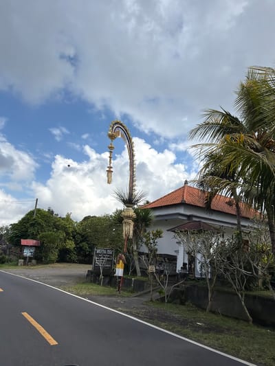 The image captures a roadside scene in Susut, Indonesia, under a bright sky with scattered clouds. A paved road with yellow and white lane markings occupies the foreground. To the right of the road is a white building with a terracotta-tiled roof, partially obscured by palm trees and bare-branched trees. A tall, ornate, golden decorative arch with hanging elements stands to the left of the building, near the road. In front of the building, two black stone signposts are visible; one has text that reads "AMERTHA YOGA" and "BALI NATURAL SPICES", while the other is partially obscured and displays text. A person wearing a yellow shirt and white pants stands near the signposts. Lush green trees and foliage surround the building and the roadside.