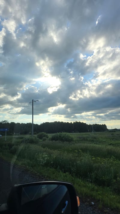 The image depicts a view from a car, looking out at a rural landscape under a cloudy sky. The sky is filled with dramatic, dark clouds interspersed with patches of bright sunlight breaking through, creating a sense of diffused light. In the foreground, the top portion of a car's side mirror is visible, reflecting the sky and a portion of the car's interior.

The landscape features a wide, green field with dense vegetation, including tall grass and scattered bushes. A line of dark green trees forms a dense forest in the middle ground. A utility pole with wires stretches across the scene. To the left, a blue road sign is visible, indicating "БОВ 7", likely a street or route number in the city of Brod, Russia. There are no people or visible activity in the scene. The overall impression is of a somewhat overcast day in a rural setting.