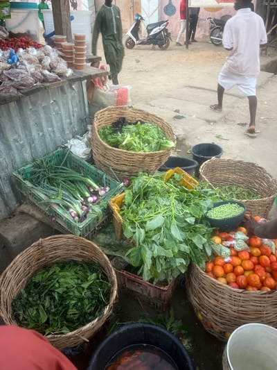 A bustling outdoor market displays a variety of produce. Baskets overflow with bright red tomatoes, green peas, lettuce, and leafy greens. Other vegetables, including green onions and possibly onions, are stacked in crates. Additional food items are visible on a nearby table. People walk through the market scene, adding to the vibrant atmosphere. A motorcycle is parked nearby.