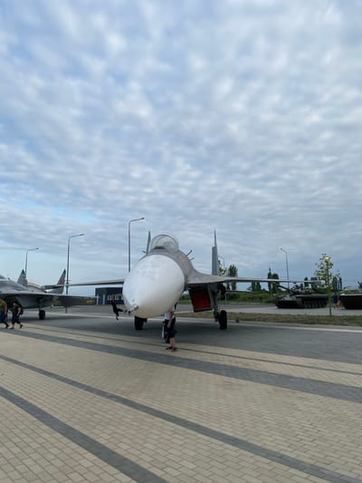 The image shows an outdoor display of military vehicles, likely a museum or exhibition. The foreground features the nose of a fighter jet with a white, pointed nose cone. Behind it, another fighter jet is visible, and further in the background, there are at least two tanks. The ground is paved with interlocking bricks in a pattern of light and dark gray. Several people are walking around the display. The sky is cloudy with a mixture of white and gray clouds. The location is identified as Arablyar, Russia.