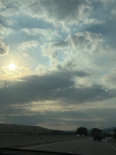 The image depicts a cloudy sky over a highway, likely in or near Yeni Qəsəbə, Azerbaijan. The sun is partially visible through the clouds on the left side of the frame, casting a soft glow. In the distance, a hilly landscape is visible, with some trees and structures suggesting a rural or suburban environment. A highway with a concrete barrier runs across the foreground, with a few vehicles on it, including a truck and some cars. The overall atmosphere is overcast, with dramatic cloud formations dominating the sky.