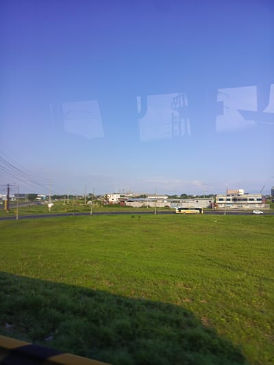 The image shows a landscape in Durán, Ecuador, captured through a window with reflections. In the foreground, there's a large grassy field, transitioning from bright green to darker shades due to shadows. A two-lane road runs behind the field with a yellow bus traveling toward the right. Several buildings are visible along the road, including a two-story structure with visible signage in the upper floor. Utility poles run along the road. The sky is clear and blue.