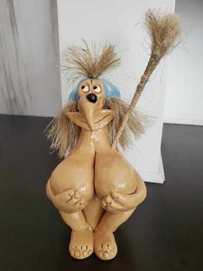 This image features a ceramic figurine, likely a whimsical representation of a dog or other creature, in a reclined position. The figurine has a light tan, glossy finish, with prominent facial features including wide, cartoonish eyes and an open mouth. It wears a bright blue head covering or cap. Strands of coarse, straw-like material, resembling hair or a mane, sprout from its head and also form a thick rope-like appendage extending from its upper body. This appendage is also made of the same straw-like material, with some loose strands fanning out. The figurine's limbs are bent and positioned in a relaxed, almost playful pose, with clearly defined hands and feet.
The background is divided horizontally into two distinct areas. The upper portion shows a white surface, possibly a wall or a large piece of paper, with a slight shadow effect. Below this is a dark, reflective surface, which could be a table or countertop. The overall lighting suggests an indoor environment, with no clear indicators of time of day or weather. There is no visible text in the image. The figurine's expression is one of surprise or exaggerated joy. The setting appears to be a domestic space, possibly a home or studio, given the casual arrangement of the objects.