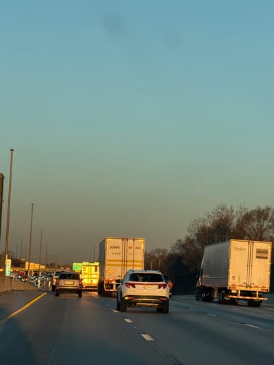 The scene depicts a multi-lane highway within the city of Hammond, United States of America, during late afternoon or early evening. The sky transitions from light blue at the zenith to an amber hue near the horizon, indicative of a low sun angle. The highway is a concrete surface with visible expansion joints and lane markings; a solid yellow line delineates the left shoulder, and white dashed lines separate traffic lanes.
Traffic is congested and moving in the same direction, away from the camera's perspective. Directly ahead in the center lane is a silver or grey SUV, bearing license plate 1AKK945. In the adjacent right lane, a white SUV with visible license plate 0JJ2664 is partially visible on the right side of the frame. Further ahead in the left lane is a white semi-trailer truck with "JOHN F. BAYARD" and "1-866-230-0807" visible on its rear. To the right of the center lane, a dark-colored semi-truck pulls a white "Trexker" trailer, displaying "NOW HIRING 618-230-1317" and "Trexker.com" on its rear. Beyond these vehicles, a yellow semi-trailer or box truck with highly illuminated rear lights is visible. Numerous other vehicles, including cars and trucks, are present in the distance, their brake lights illuminated.
Tall, unlit metal light poles line the left side of the highway. A green overhead road sign is partially visible in the distance on the left, displaying "SOUTH Danville" and another destination. The right side of the highway features a dark sound barrier or fence, behind which appear trees with sparse, bare branches, suggesting a late fall or winter season. The upper portion of the sky exhibits minor lens flares or sensor dust spots. No individual people are distinctly visible.