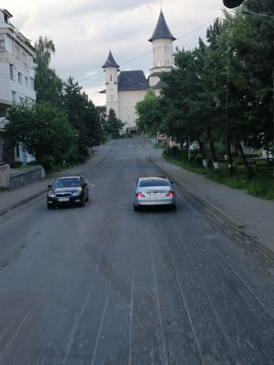 The image captures a view from a vehicle traveling along a paved road in Fălticeni, Romania. The road is lined with trees and buildings, and a large church with a prominent steeple is visible in the background. Two cars are present on the road: a black Skoda Octavia in the left lane and a silver Mercedes-Benz CLS in the right lane. The Skoda's license plate reads "SV 18 SFS", and the Mercedes' license plate is "SV 77 JJJ". The sky is overcast, and there is a noticeable amount of dust or debris on the windshield of the vehicle from which the photo was taken. The scene depicts a typical urban street in Fălticeni, likely during daylight hours.