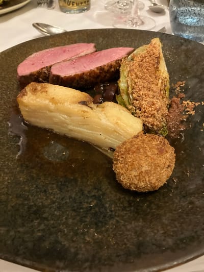 The image depicts a plate of food, possibly in a restaurant setting in Glenridding, United Kingdom. The plate contains sliced, medium-rare duck breast, a piece of breaded cabbage, a golden-brown croquette, and a pale-colored vegetable, possibly a parsnip or celery root. There are also small dark specks that could be berries or a sauce component. The plate itself is dark and textured. The lighting suggests an indoor setting, with other tableware visible in the periphery.