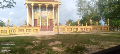 This media file captures a view of a Buddhist temple in Cambodia, with a prominent golden fence in the foreground. The temple itself is richly decorated with gold accents, intricate carvings, and murals depicting Buddhist scenes. The central facade features two large doors, with a grand mural above them showing figures in a serene setting, likely related to the Buddha's life or teachings. Flanking this are pillars adorned with golden motifs. The fence, also in a striking golden hue, runs across the middle ground and is adorned with decorative balusters and spires. Beyond the fence and temple, lush green trees dominate the background, suggesting a natural and somewhat rural setting. The sky is overcast, indicating a cloudy or possibly rainy day. In the lower left corner of the image, a timestamp and text are visible. The timestamp reads "2026/06/18 16:00," and the text below it appears to be in Khmer script, followed by the "TECNO" logo. The overall atmosphere is one of cultural and spiritual significance, set against a backdrop of natural greenery under a muted sky. No people are clearly visible, and the scene appears to be a static depiction of the temple and its surroundings. A small dog is visible on the stairs of the temple.